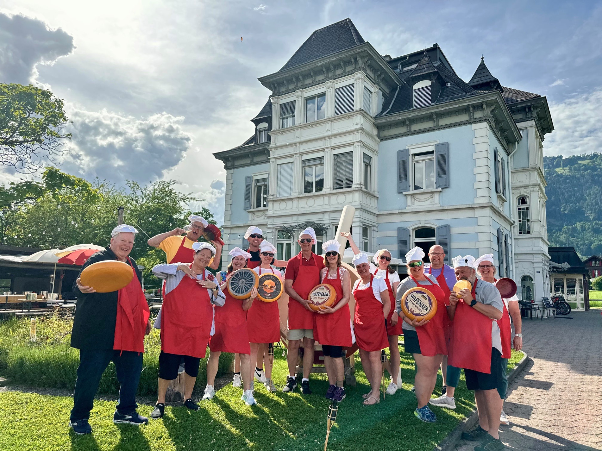 A group of Journey with Erika travelers in red aprons and chef hats holding cheese wheels outside a Swiss mansion